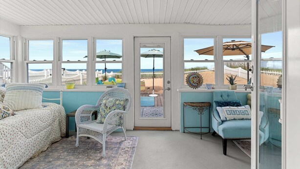 A sunlit room with a view of the beach, featuring a bed, two chairs, and a small table.
