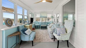 A spacious room with a ceiling fan, large windows, a TV, and a cozy seating area with patterned cushions.
