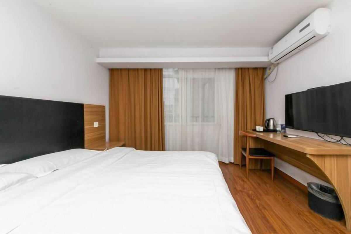 A hotel room with a bed, a desk, a chair, a TV, and an air conditioning unit.