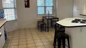 Una cocina con una isla central, dos taburetes de bar, una mesa de comedor y una lámpara colgante.