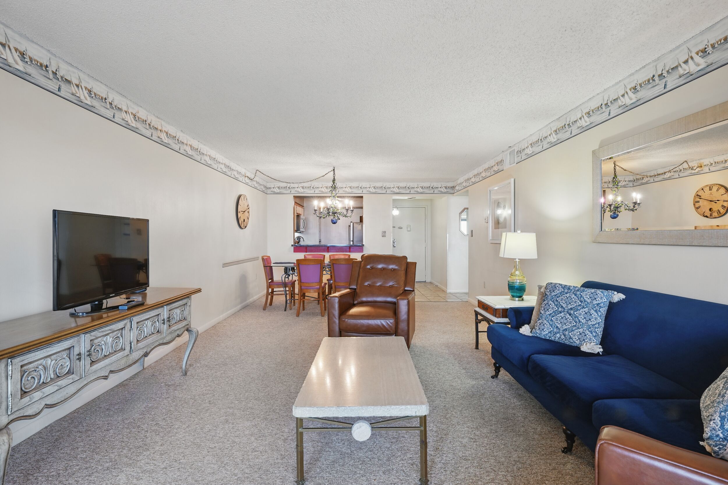 A living room with a TV, sofa, coffee table, and dining area with chairs and a chandelier.
