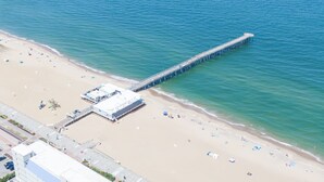 An aerial view of a beach with a long pier extending into the sea, a sandy beach with umbrellas, and a building with a flat roof near the shore. - The Colony at Virginia Beach (Virginia Beach)