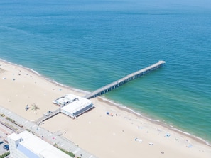 An aerial view of a beach with a long pier extending into the sea, a sandy beach with umbrellas, and a building with a flat roof near the shore. - The Colony at Virginia Beach (Virginia Beach)