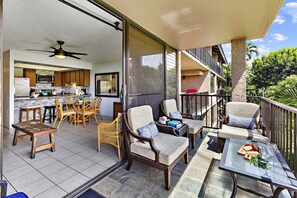 A covered outdoor patio with wicker furniture, a glass table, and a view of a kitchen and balcony.