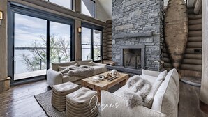 A spacious living room with a stone fireplace, large windows, and a view of a lake. - 9 bed 9 bath log home on Lake Muskoka (Bracebridge)