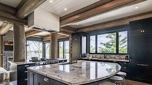 A modern kitchen with a large island, dark cabinetry, and a stone fireplace. - 9 bed 9 bath log home on Lake Muskoka (Bracebridge)