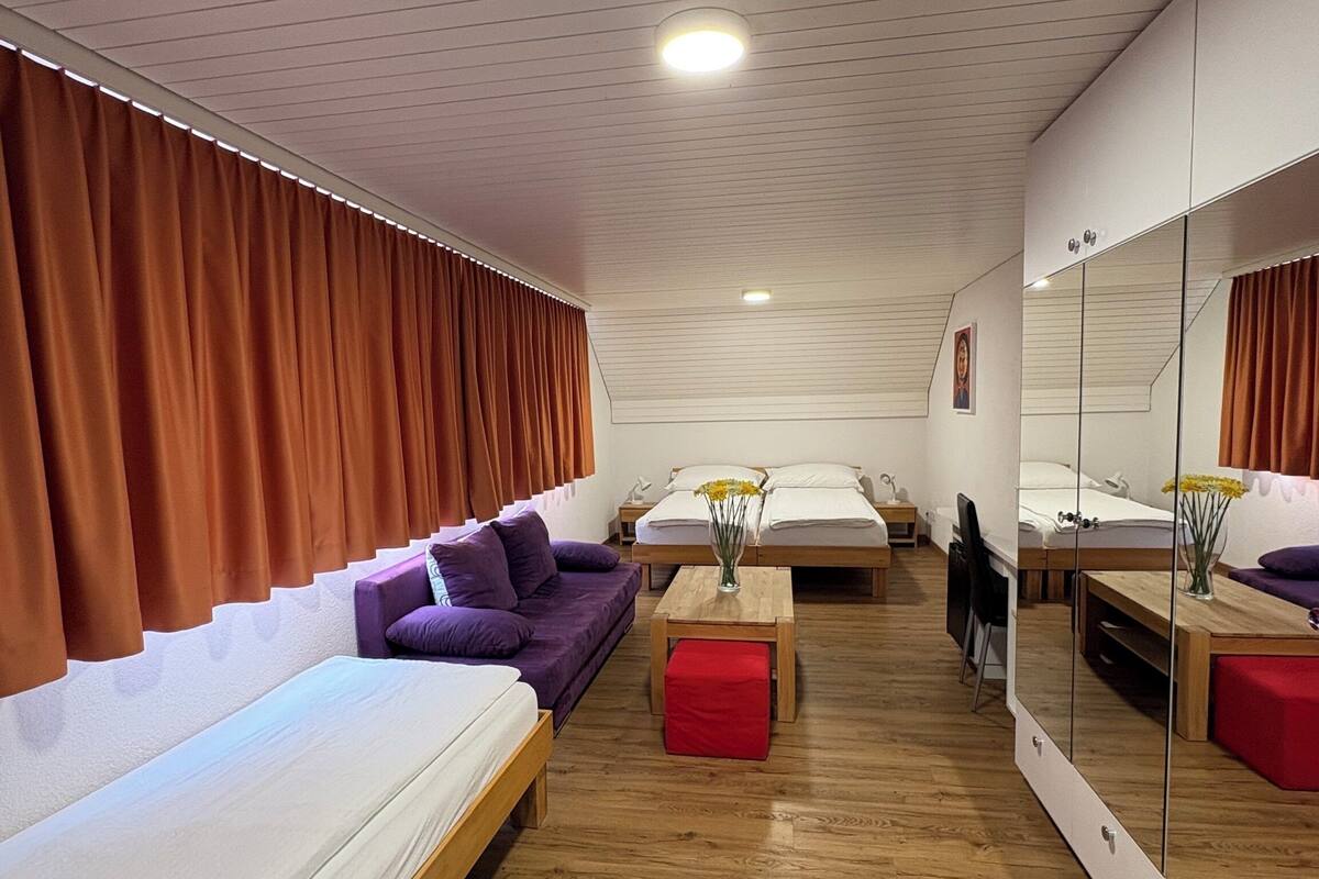 Une chambre d’hôtel moderne avec un plancher en bois, un canapé violet, un lit, une petite table avec un vase et un grand miroir.