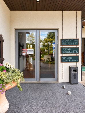 Una entrada de edificio con puerta de vidrio, un letrero publicitando una hamburguesa con piedras calientes, y un cartel de bienvenida.