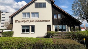 A two-story building with a sign reading "Wirtschaft zum Himmel". It has multiple windows, a sloped roof, and is surrounded by greenery.