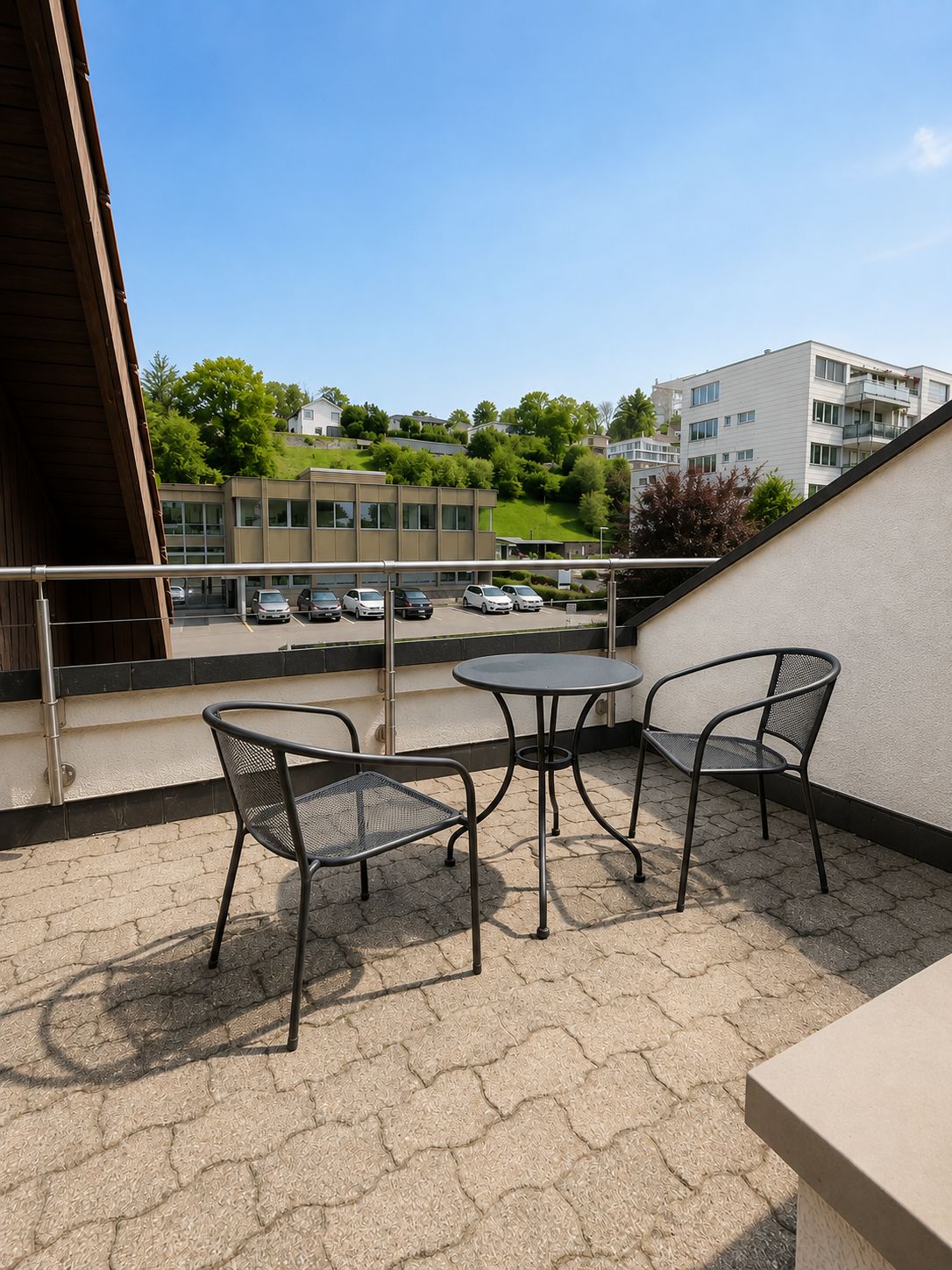 A balcony with two metal chairs and a small round table, overlooking a parking lot and buildings.