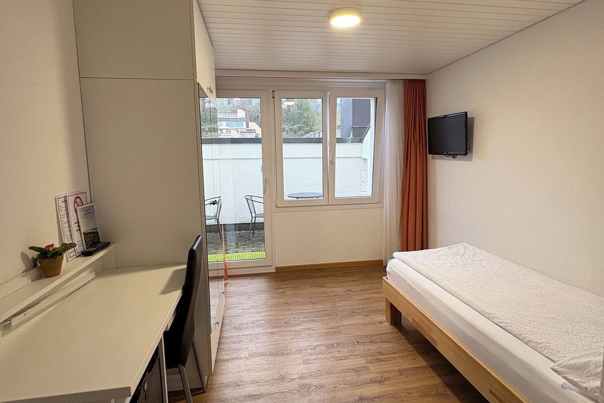 Une petite pièce comprenant un lit, un bureau, une chaise, un téléviseur et une fenêtre avec des rideaux.