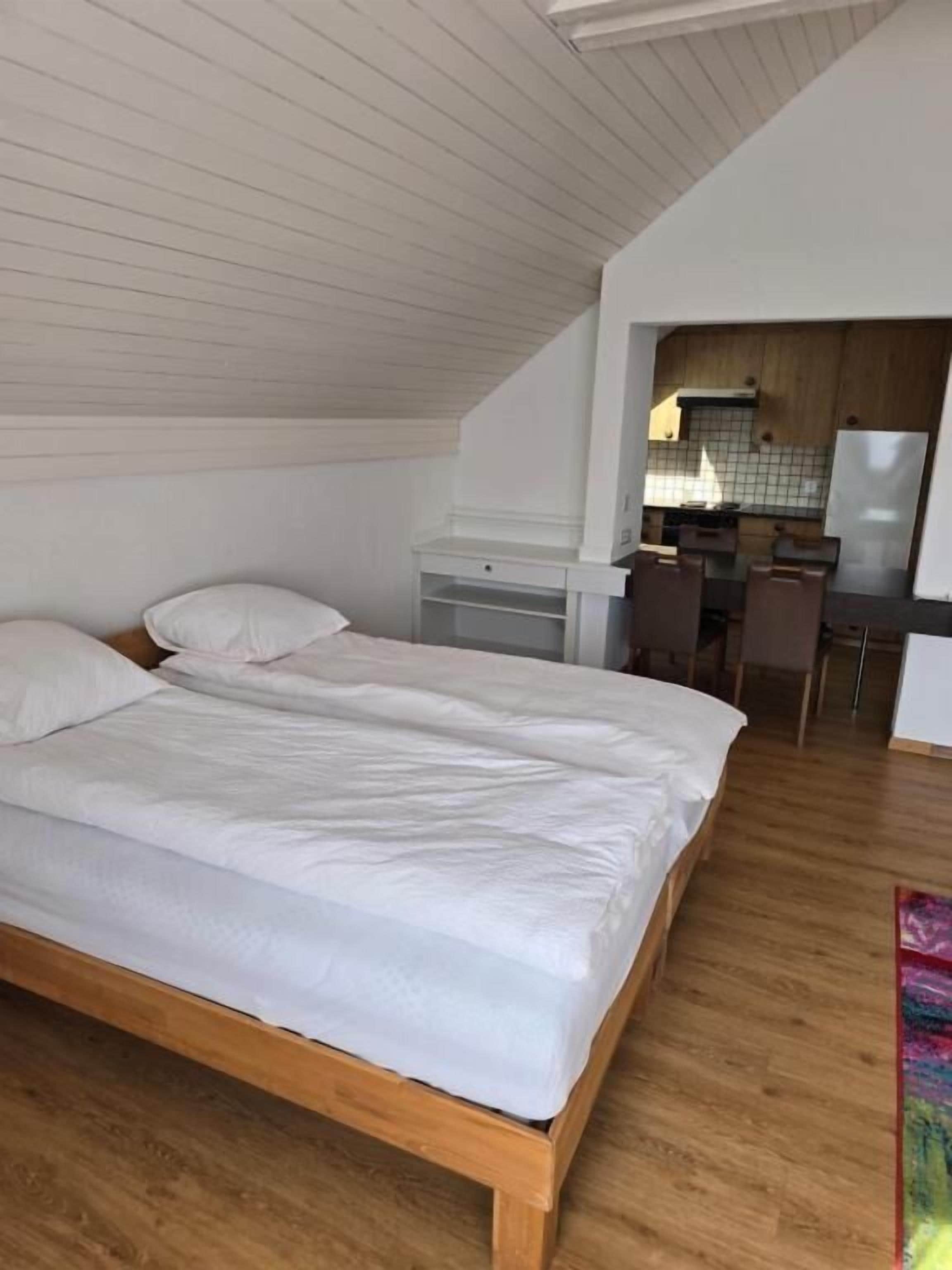 A bedroom with a wooden bed, a desk, and a kitchenette in the background.