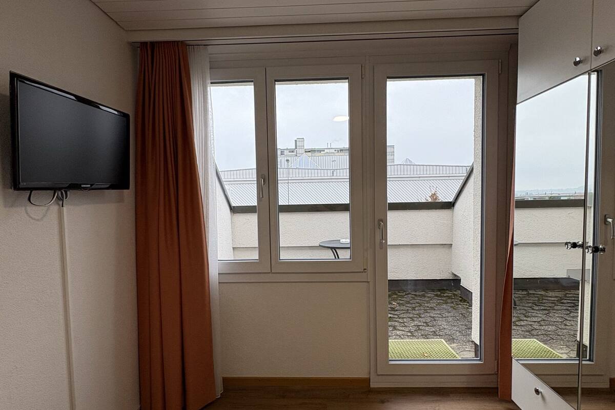 Une chambre avec un plancher en bois, un lit, un téléviseur fixé au mur et une fenêtre donnant sur un immeuble et un jardin.