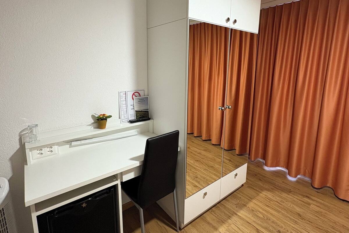 Une pièce de taille modeste, avec un bureau blanc, une chaise, une petite plante, un miroir et une armoire aux portes coulissantes.