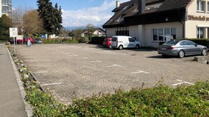 Un estacionamiento con varios coches, un edificio con un cartel que dice "Wirtschaft" y un árbol grande al fondo.
