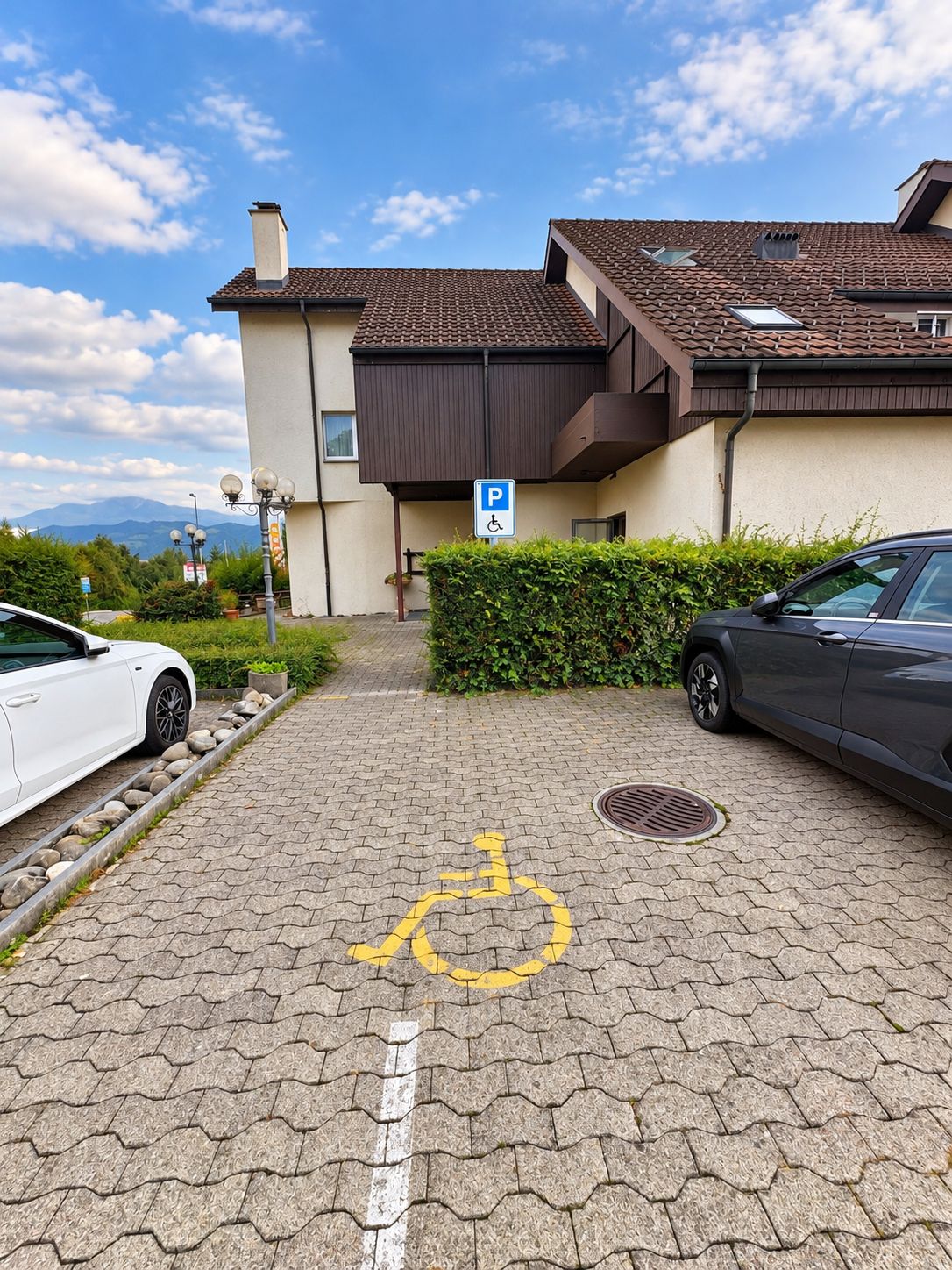 Une zone de stationnement avec un panneau indiquant les places réservées aux personnes handicapées, un symbole de fauteuil roulant peint au sol, et un bâtiment avec un toit brun et des murs de couleur beige.