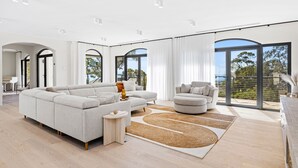 Ein geräumiges Wohnzimmer mit einer großen Eckcouch, einer kleineren Couch und einem Couchtisch. Große Fenster mit Vorhängen, Blick ins Freie und Holzboden.