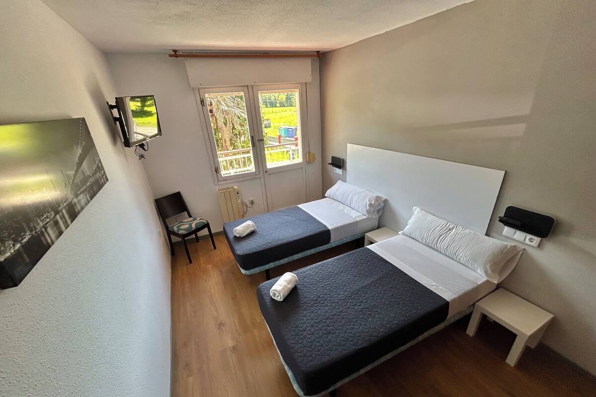 A hotel room with two beds, a small table, a chair, and a TV mounted on the wall.