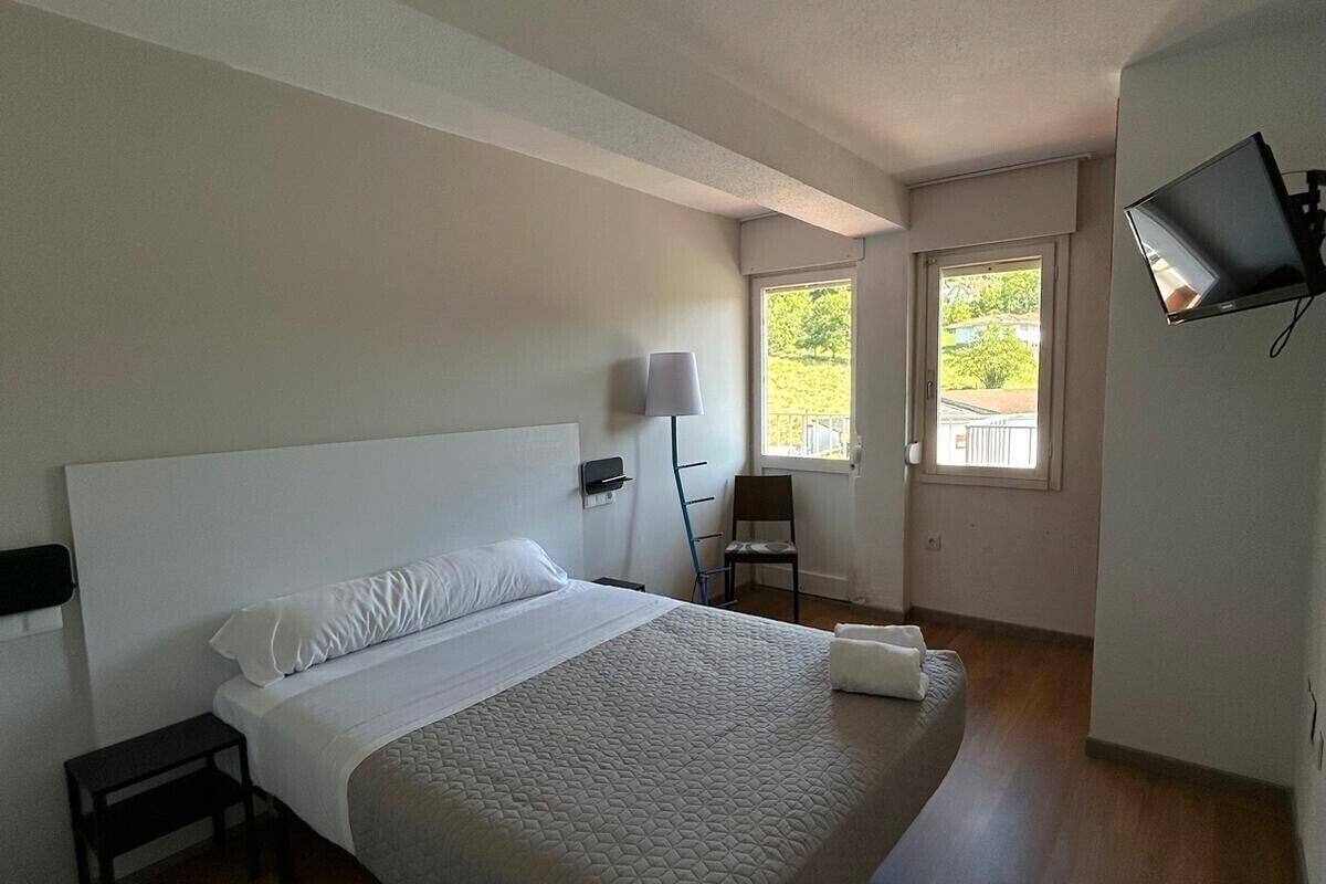 A bedroom with a bed, a nightstand, a chair, a TV, and a window.