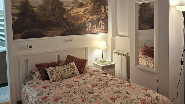 A bedroom with a bed, a nightstand, a lamp, a mirror, and a large landscape painting on the wall.