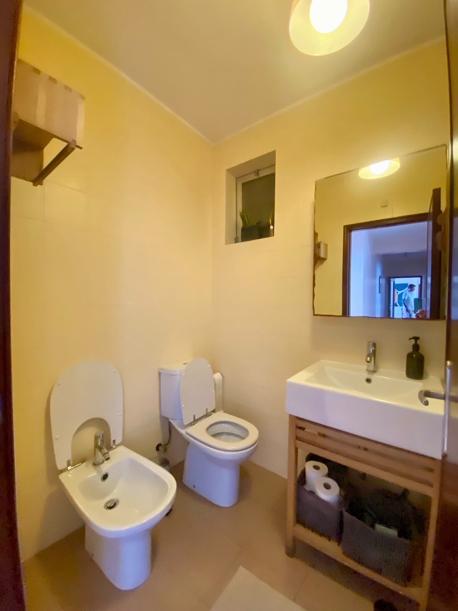 A bathroom with two toilets, a sink, a mirror, and a shelf with toiletries.