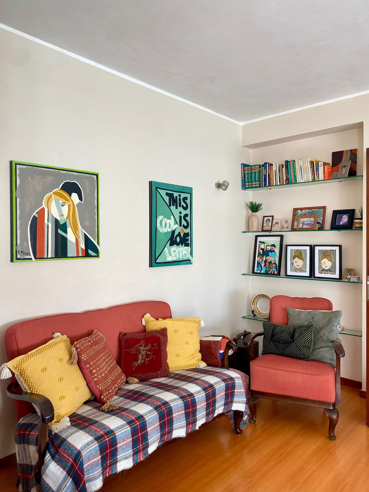 Ein Wohnzimmer mit einem roten Sofa, einer karierten Decke und einem roten Sessel. An der Wand hängen zwei gerahmte Kunstwerke und ein Bücherregal mit Büchern und Dekorationen.