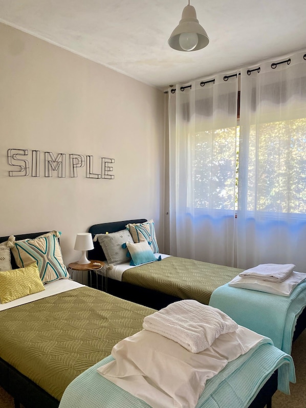 A room with two beds, a wall-mounted lamp, and a window with sheer curtains.