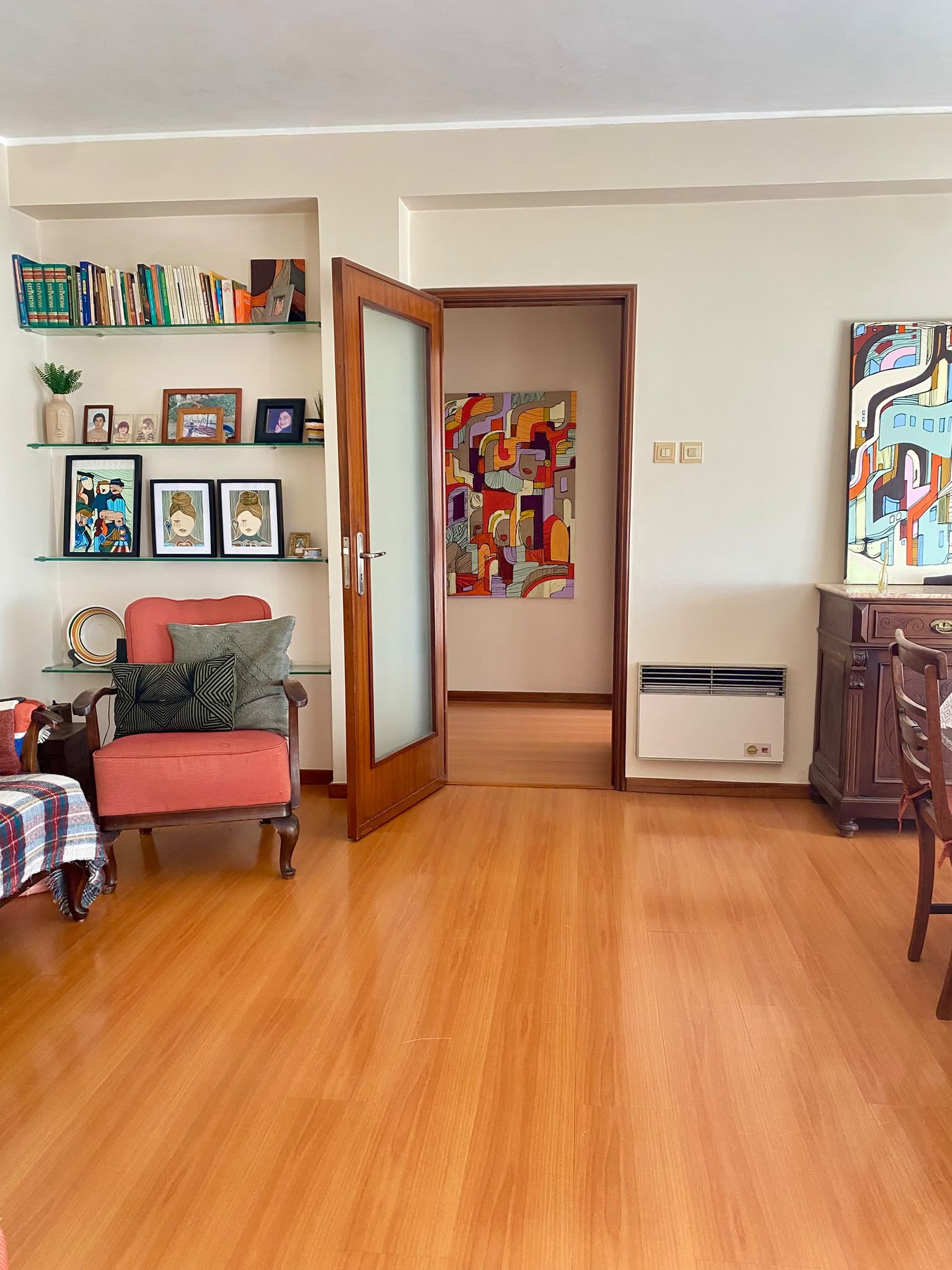 Un salon avec un canapé rouge, une chaise en bois, une bibliothèque et un tableau abstrait aux couleurs vives.