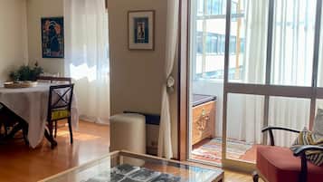 Un salon avec une table basse à plateau en verre, un coin repas avec une table et des chaises, et une grande fenêtre avec des rideaux.