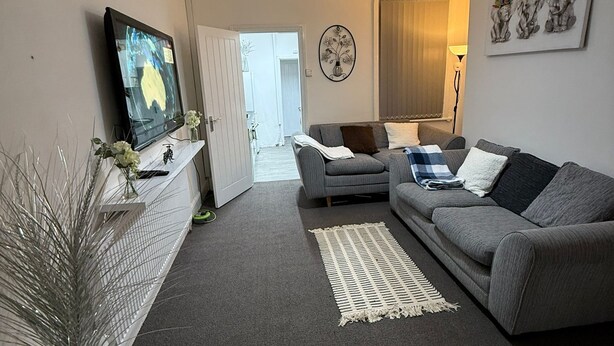A living room with a grey sofa, a TV on the wall, a white vase with plants, and a door leading to another room.