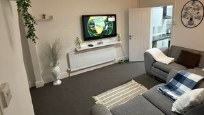 A modern living room with a grey sofa, a television on a white stand, and a kitchen visible through an open door.