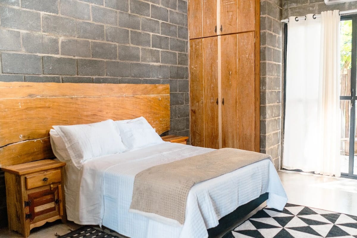 A bedroom with a wooden headboard, a bed with white bedding, a small wooden nightstand, and a large window with a view of the outside.