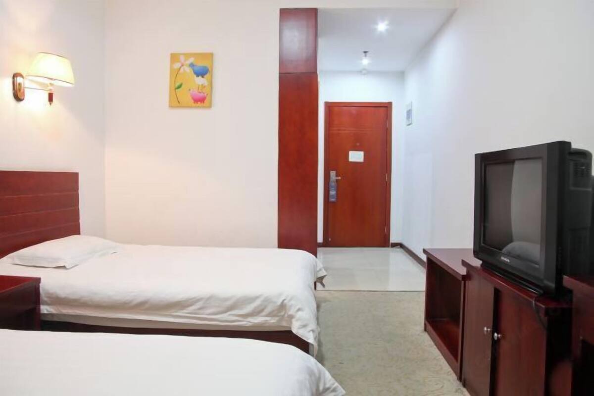 Ein Hotelzimmer mit zwei Einzelbetten, einem Fernseher, einem Holzschrank, einer Tür und einer Wandlampe.