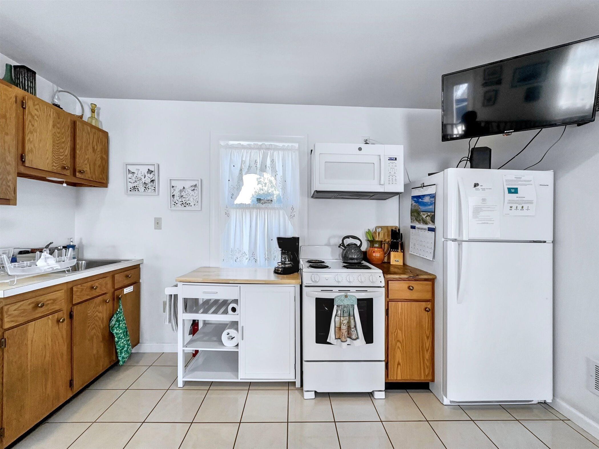 A compact kitchen with wooden cabinets, a white countertop, a stove, a microwave, a refrigerator, and a television mounted on the wall.