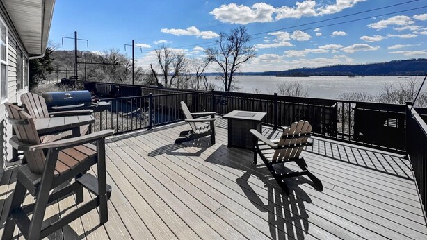 Eine Terrasse mit Gartenmöbeln, einem Grill und Blick auf einen See und Berge.