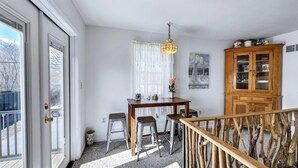 Ein Essbereich mit einem Holztisch, zwei Metallhockern, einer hängenden Leuchte und einem Holzschrank mit Glastüren.