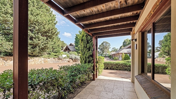 A covered outdoor area with a stone floor, surrounded by greenery and a view of a building with a sloped roof.