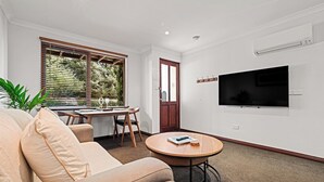 A living room with a sofa, a round coffee table, a TV mounted on the wall, and a dining area with a table and chairs.