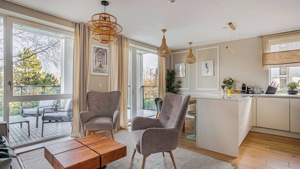 Een moderne woonkamer met een groot raam, twee grijze fauteuils, een houten salontafel, en een keukenhoek met een eettafel en een bar.