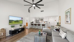 A modern living room with a flat-screen TV, a ceiling fan, a sofa, a coffee table, and a kitchen area in the background. - Brand new fully remodeled house in Palm Desert w/ golf course views-Pet Friendly (Palm Desert)