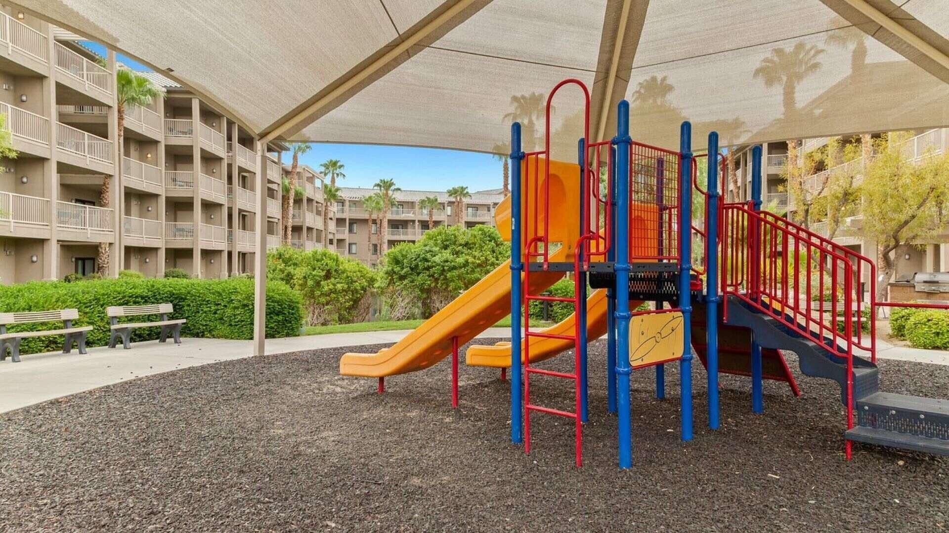 A playground with a slide and climbing structures, surrounded by a covered area and a building complex in the background.