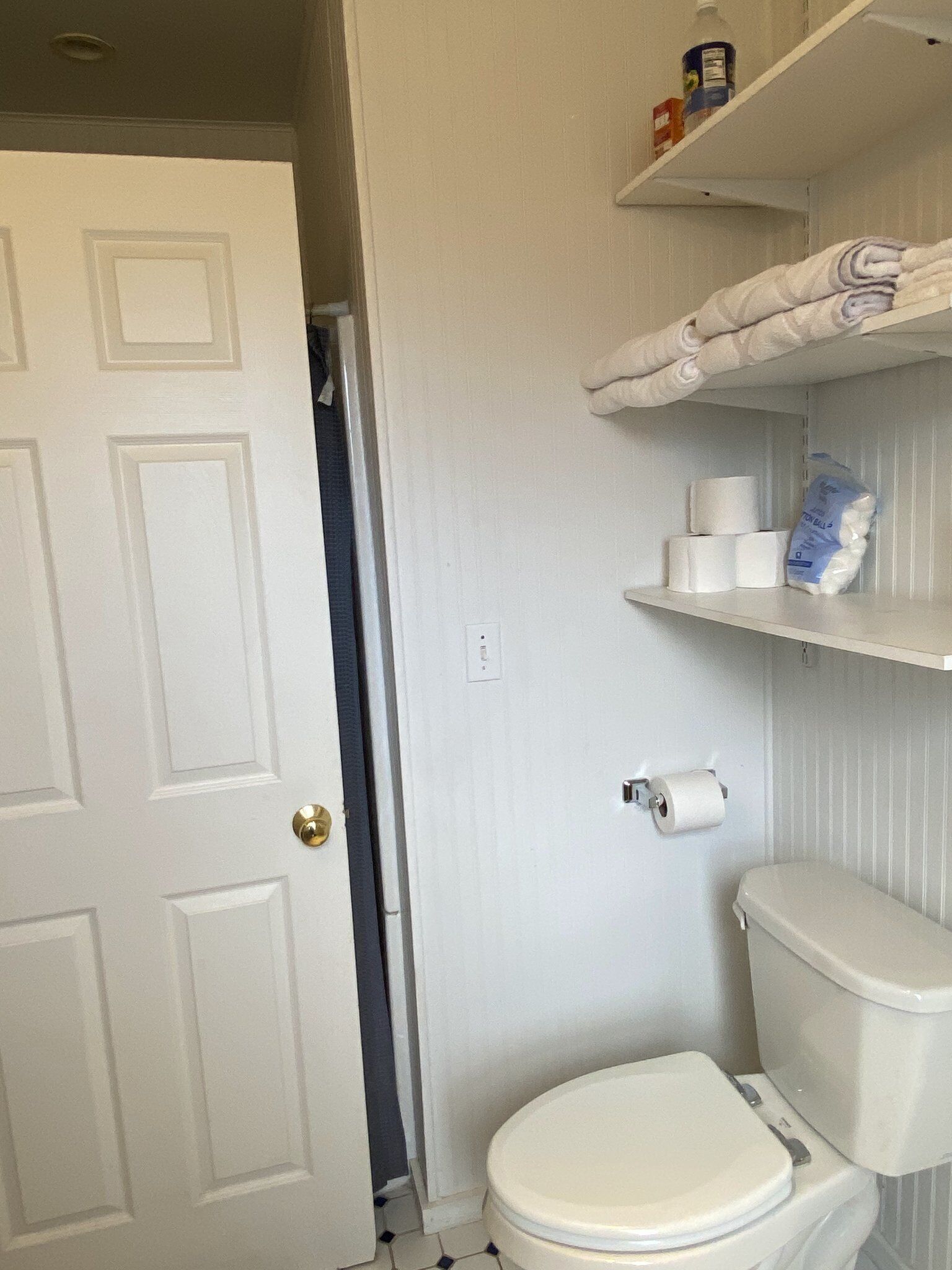 A bathroom with a toilet, a roll of toilet paper, and a shelf with towels and a bottle.