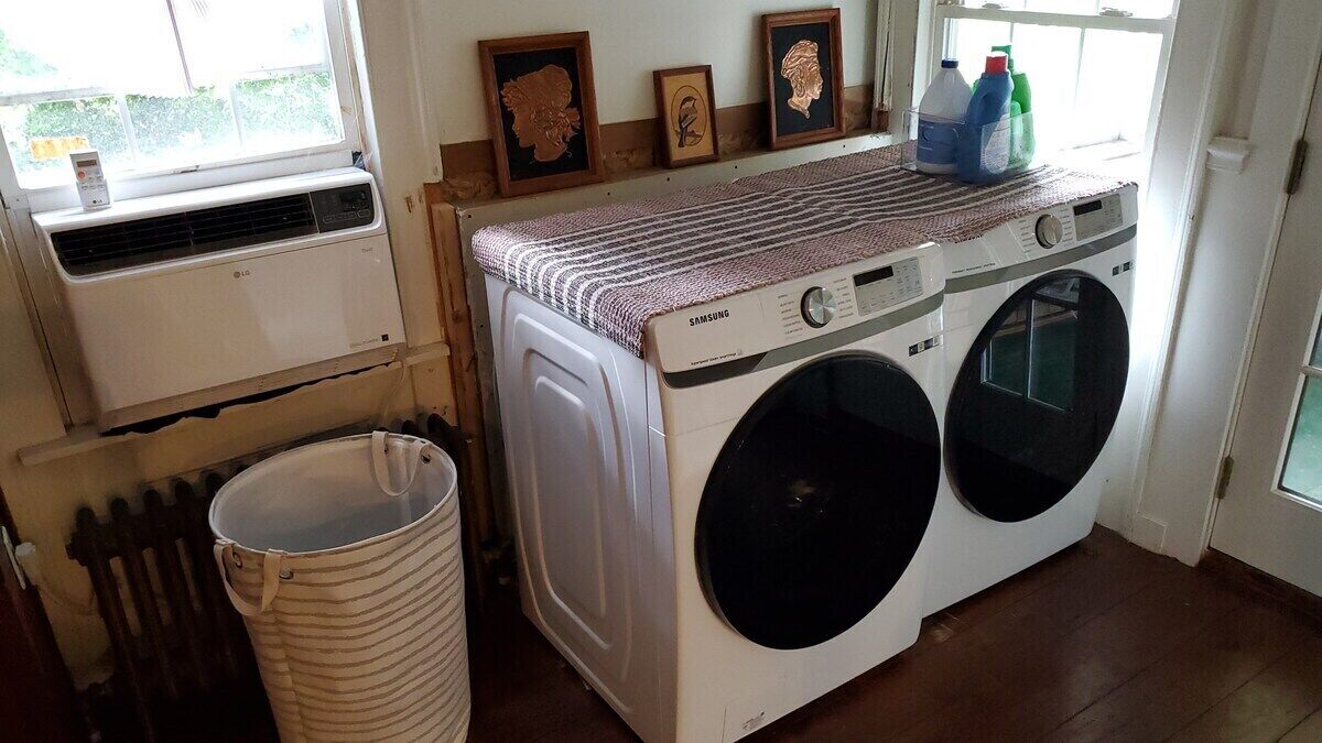 Une buanderie équipée d’un lave-linge et d’un sèche-linge, d’un radiateur, d’une poubelle et de tableaux encadrés accrochés au mur.