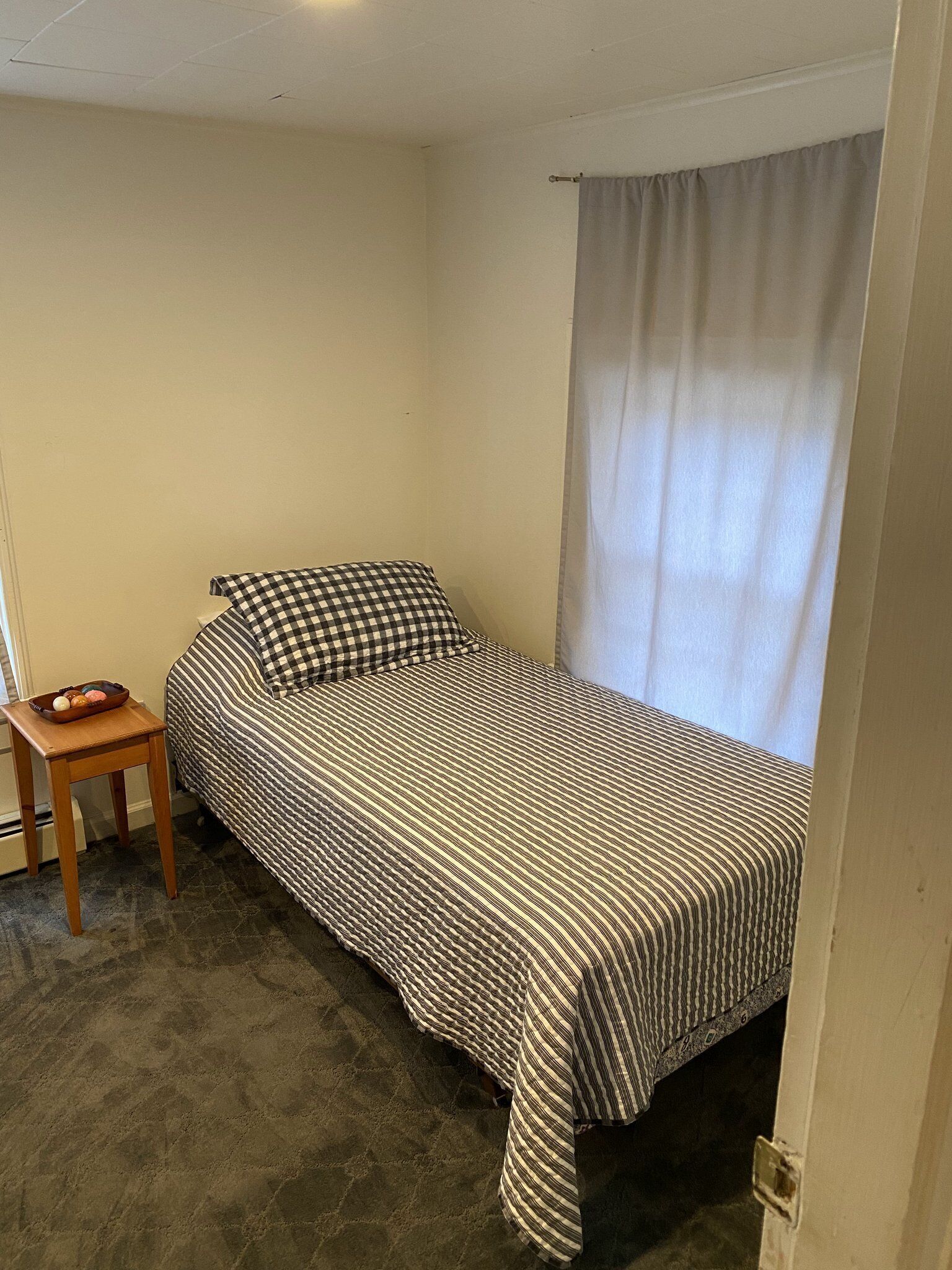 Une petite chambre avec un lit simple, une couverture à carreaux, une table de chevet en bois et une fenêtre ornée d’un rideau blanc.