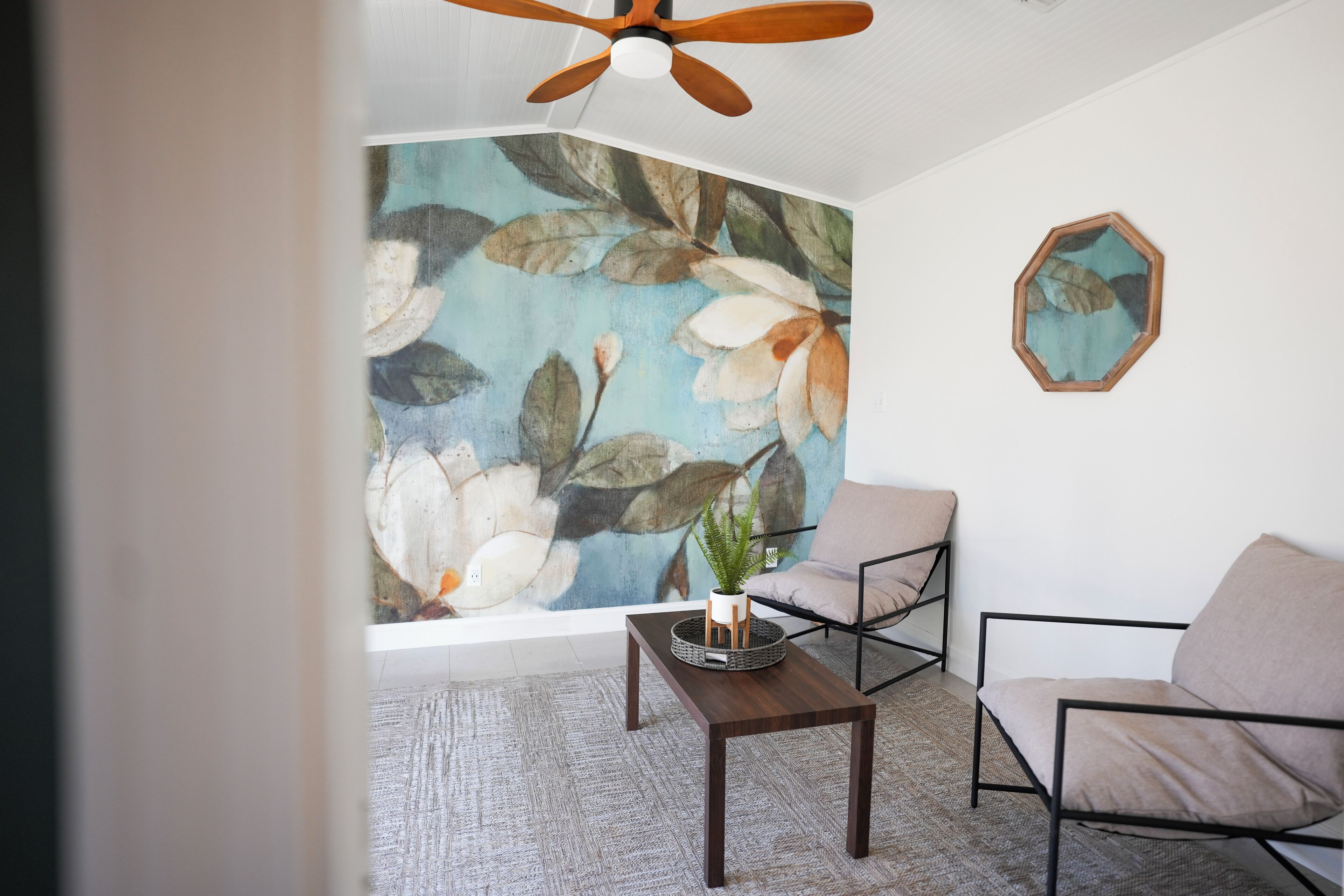 A room with a large floral mural, a ceiling fan, a modern chair, a small table with a plant, and a decorative mirror.