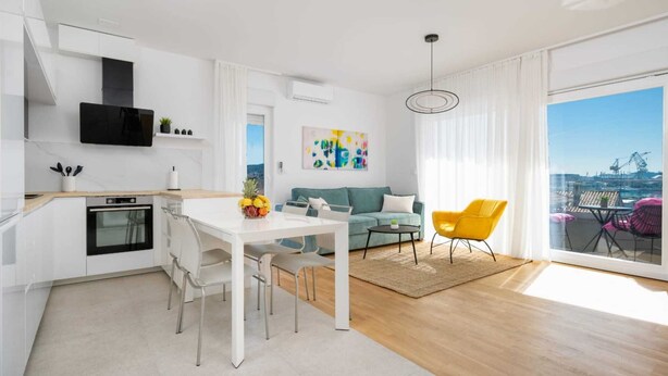 A modern apartment with a kitchen, dining area, and living room. It features a large window with a city view, a dining table with chairs, a sofa, and a yellow armchair. - Seaview apartment Lana (Seget Donji)