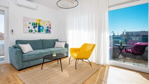A modern living room with a teal sofa, a yellow armchair, a small coffee table, and a large window with a view of the city. - Seaview apartment Lana (Seget Donji)