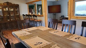 A dining area with a wooden table set for six, surrounded by chairs. A wooden cabinet with glass doors is to the left, and a television is mounted on the wall to the right. - Hale Eddy (Hancock)