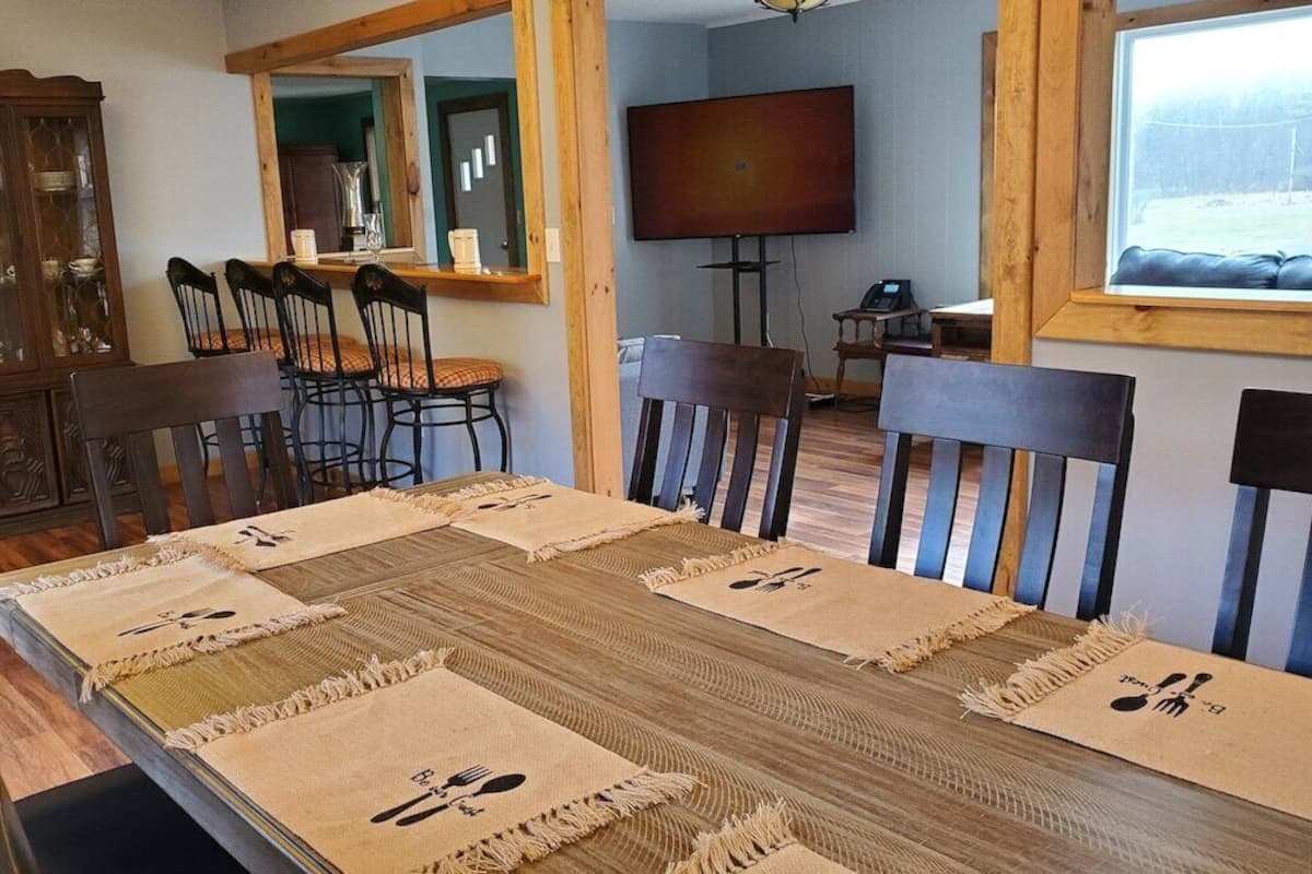 A dining area with a wooden table set for six, surrounded by chairs. A wooden cabinet with glass doors is to the left, and a television is mounted on the wall to the right.
