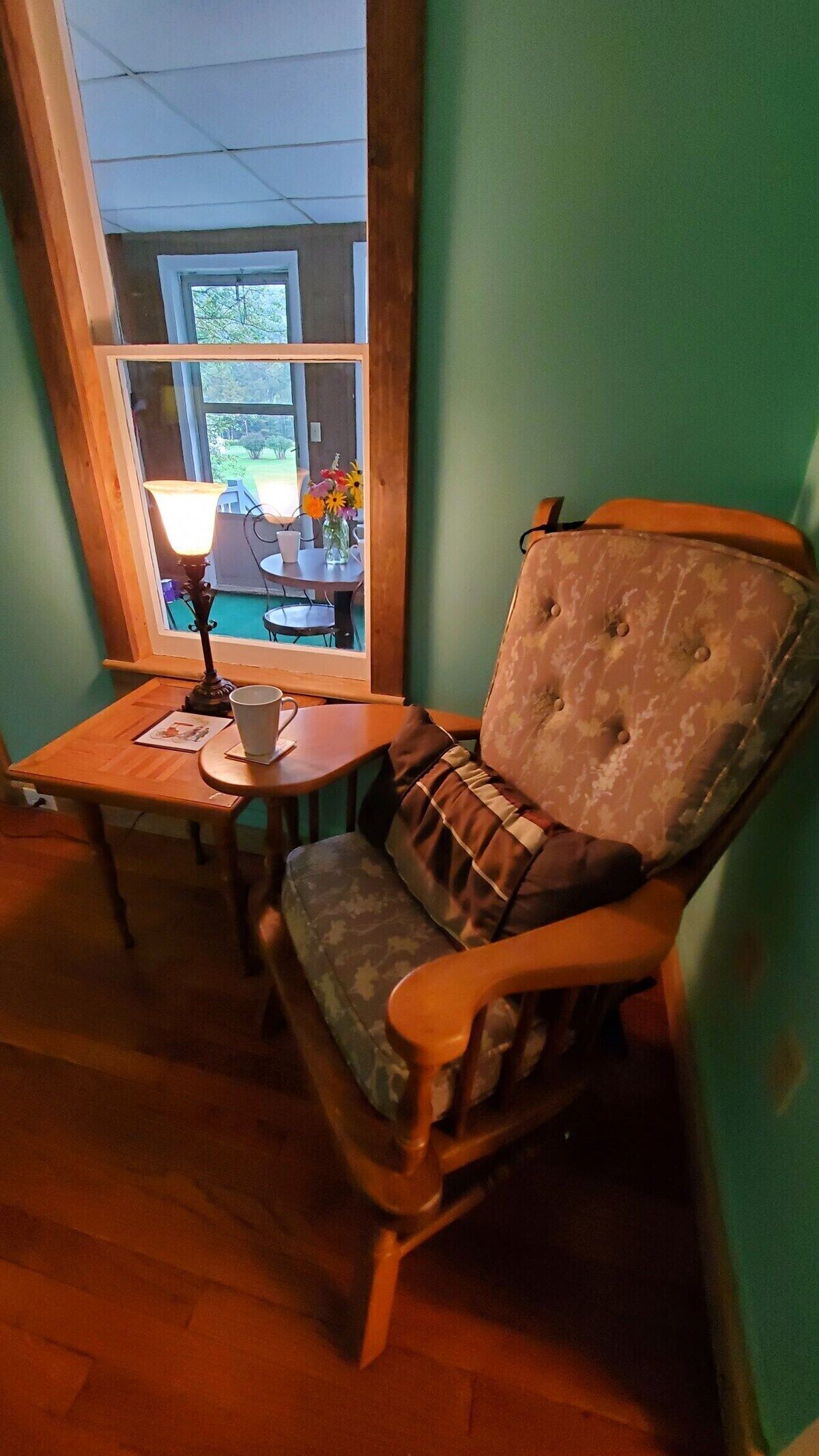 A cozy room with a wooden chair, a small table, and a window overlooking a garden.